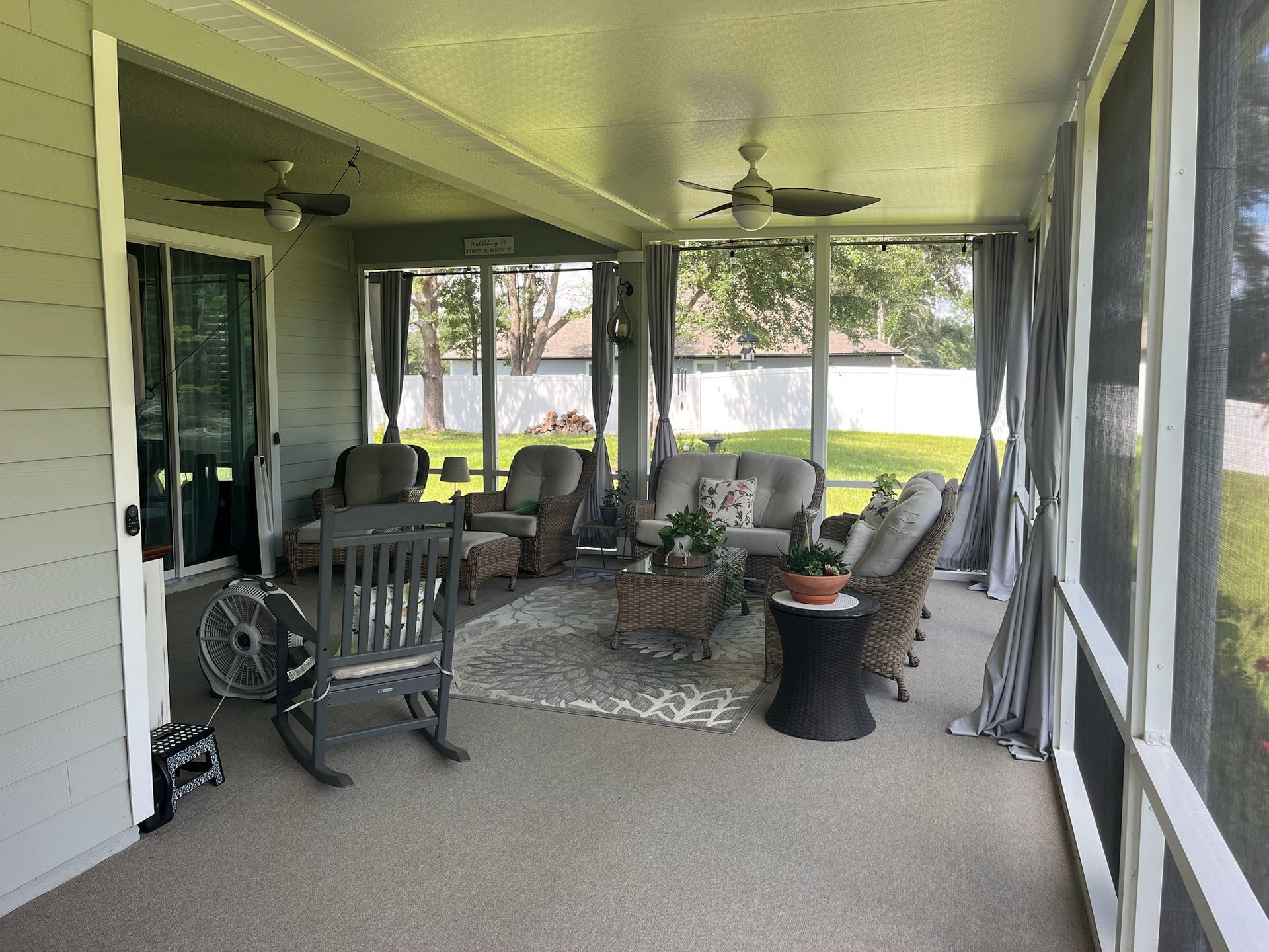 Screened-in porch with seating, rug, and ceiling fans. Overlooking a yard with trees.