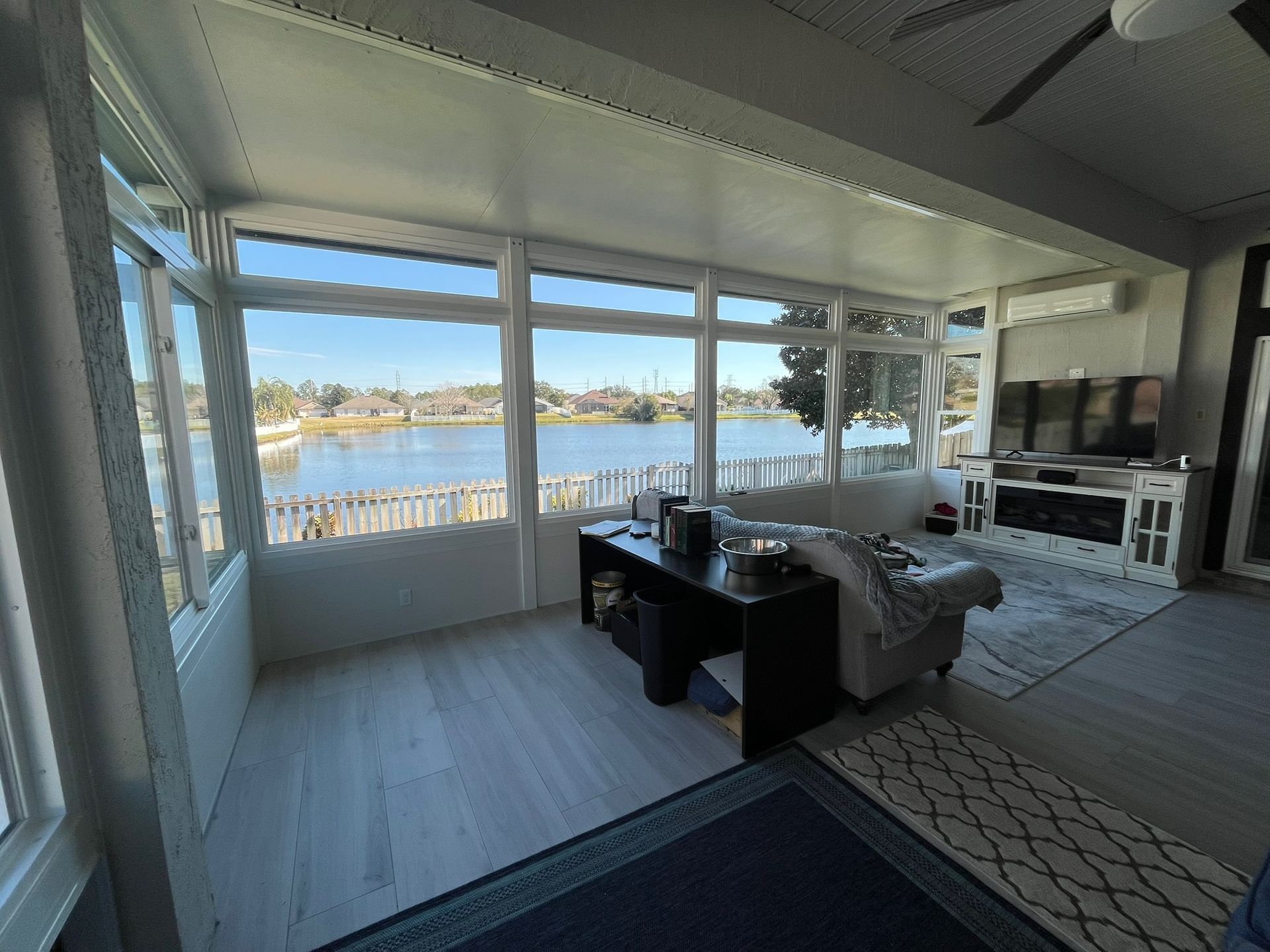 Sunroom with large windows overlooking a lake. Gray floors, white trim, dark furniture, and a blue rug.