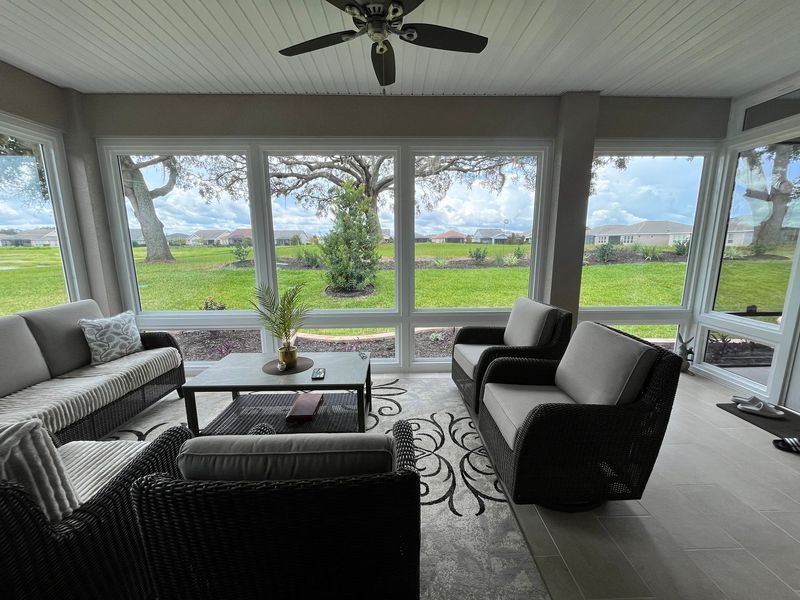 Sunroom interior with outdoor view. Gray sofas, coffee table, rug, ceiling fan, and large windows.