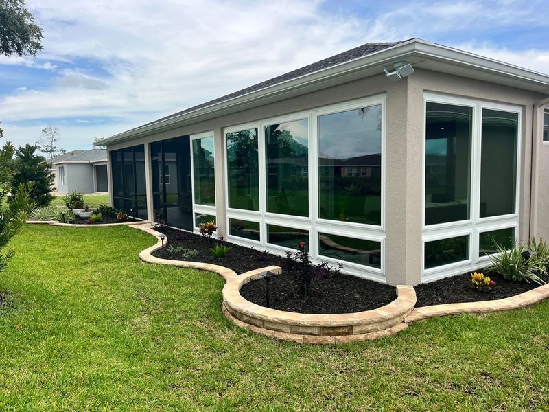 Sunroom with large windows and a screened door, beige exterior, and a small garden with a stone border.