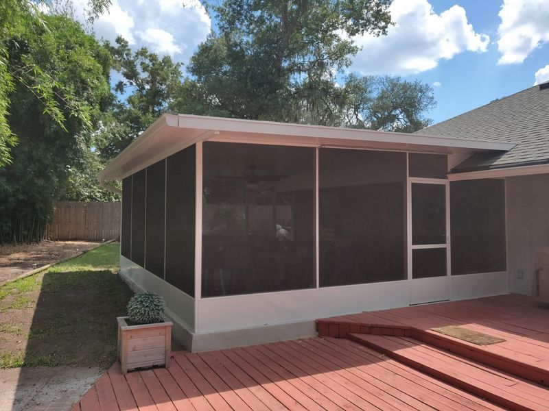 Screened-in porch attached to a house with a red wooden deck, overlooking a yard with trees and a light blue sky.