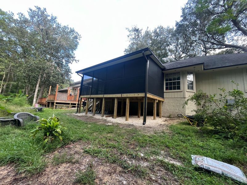 Screened-in porch attached to a light-colored house. Dark screen, wooden supports, and grassy yard.