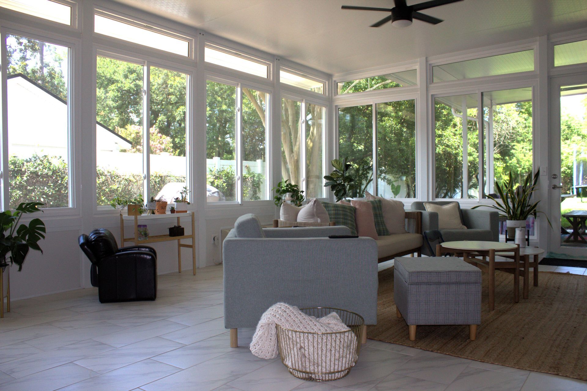 Sunroom with white walls, large windows, and light blue sofas. Basket, plants, and side tables create a cozy atmosphere.