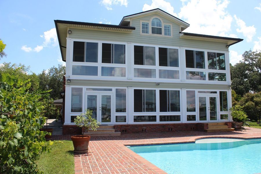 Two-story house with many windows and a pool. Exterior is light green with a red brick patio. Sunny day.