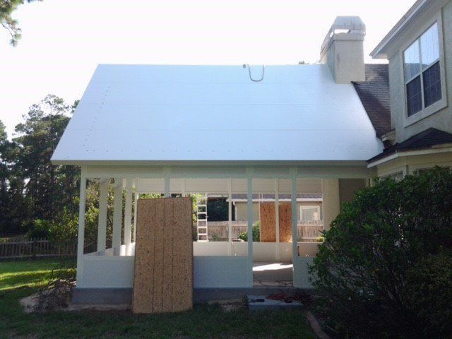 White-roofed screened porch under construction attached to a house with a chimney and windows, with plywood leaning against the porch.