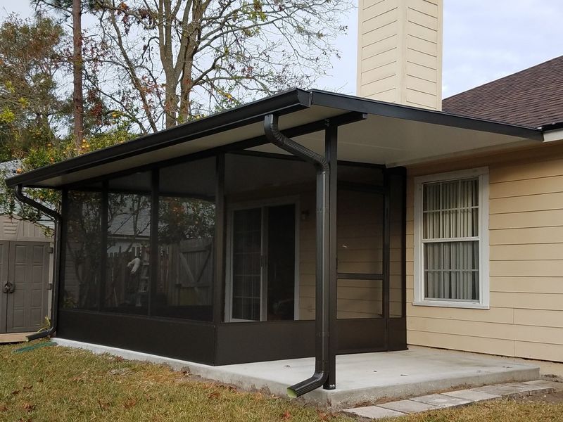 Screened-in porch attached to a tan house with a dark roof and gutters; concrete patio, and a shed in the background.