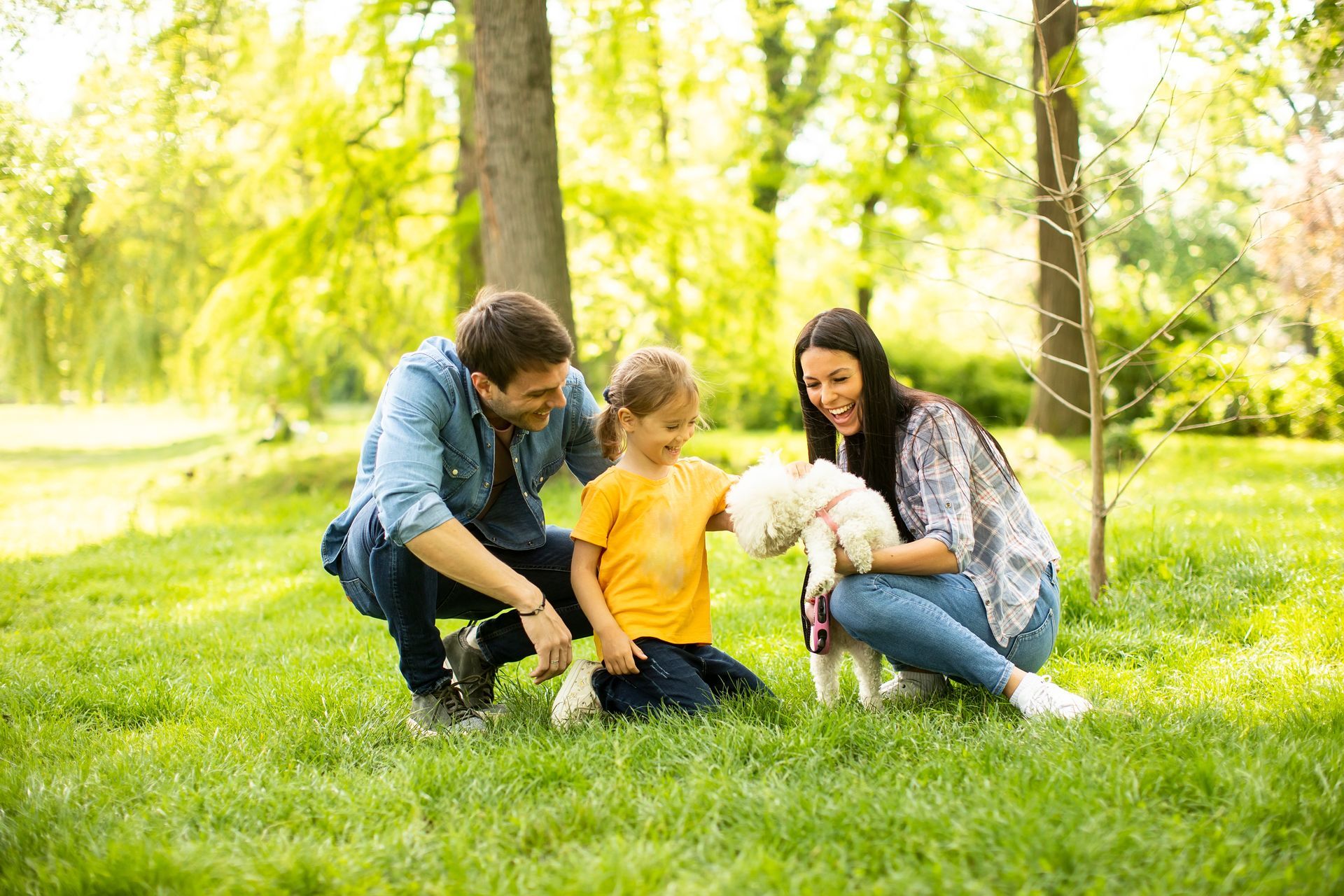 Family kneeling on grass outdoors, smiling with a white dog in a sunny park