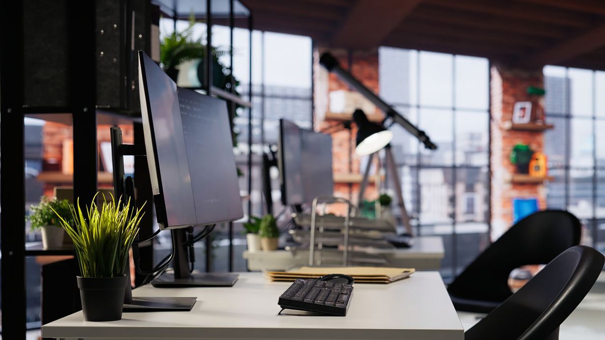 Modern office desk with computer monitors, keyboard, plants, and blurred people in the background