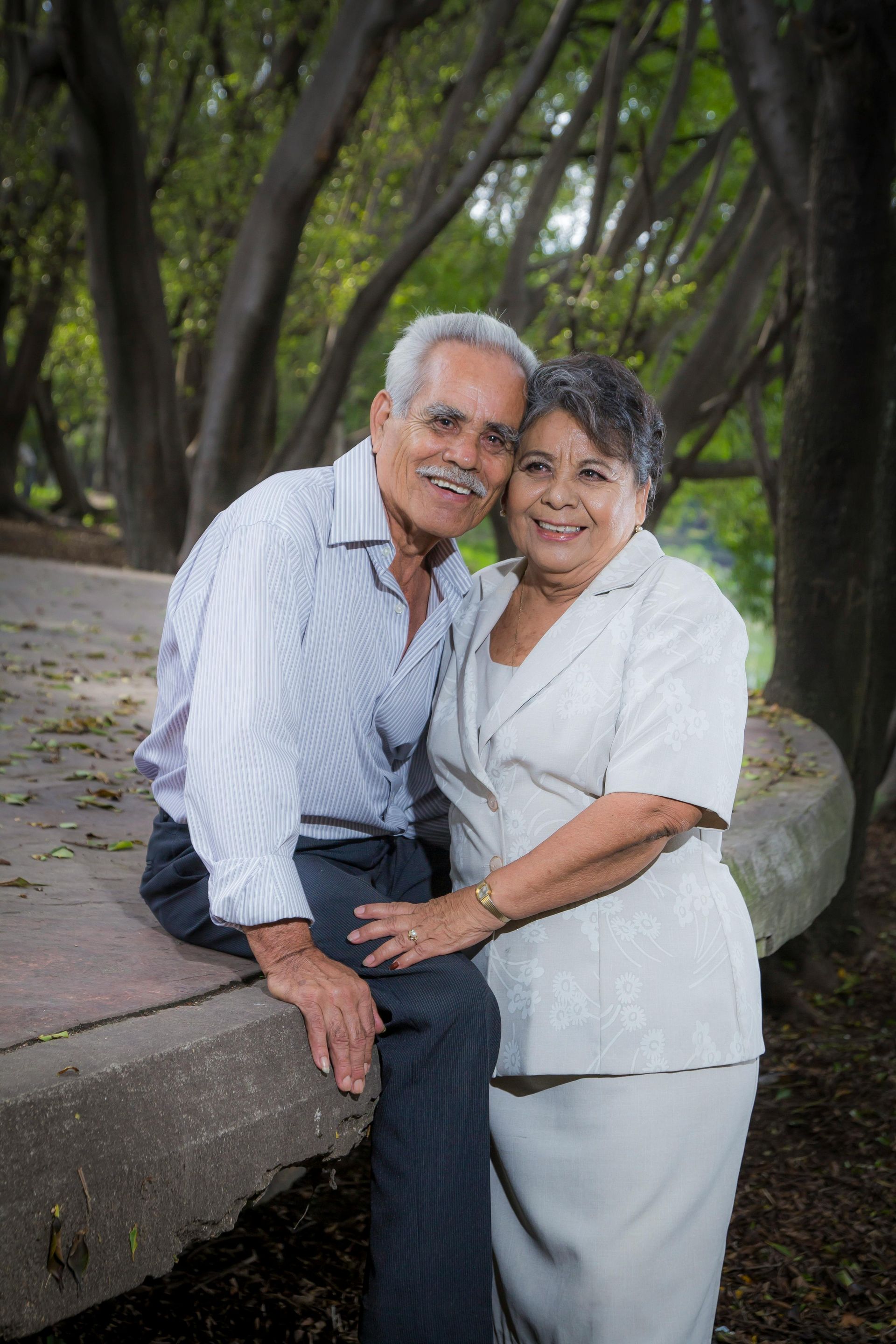Two people smiling outdoors by a stone bench, surrounded by trees and greenery.