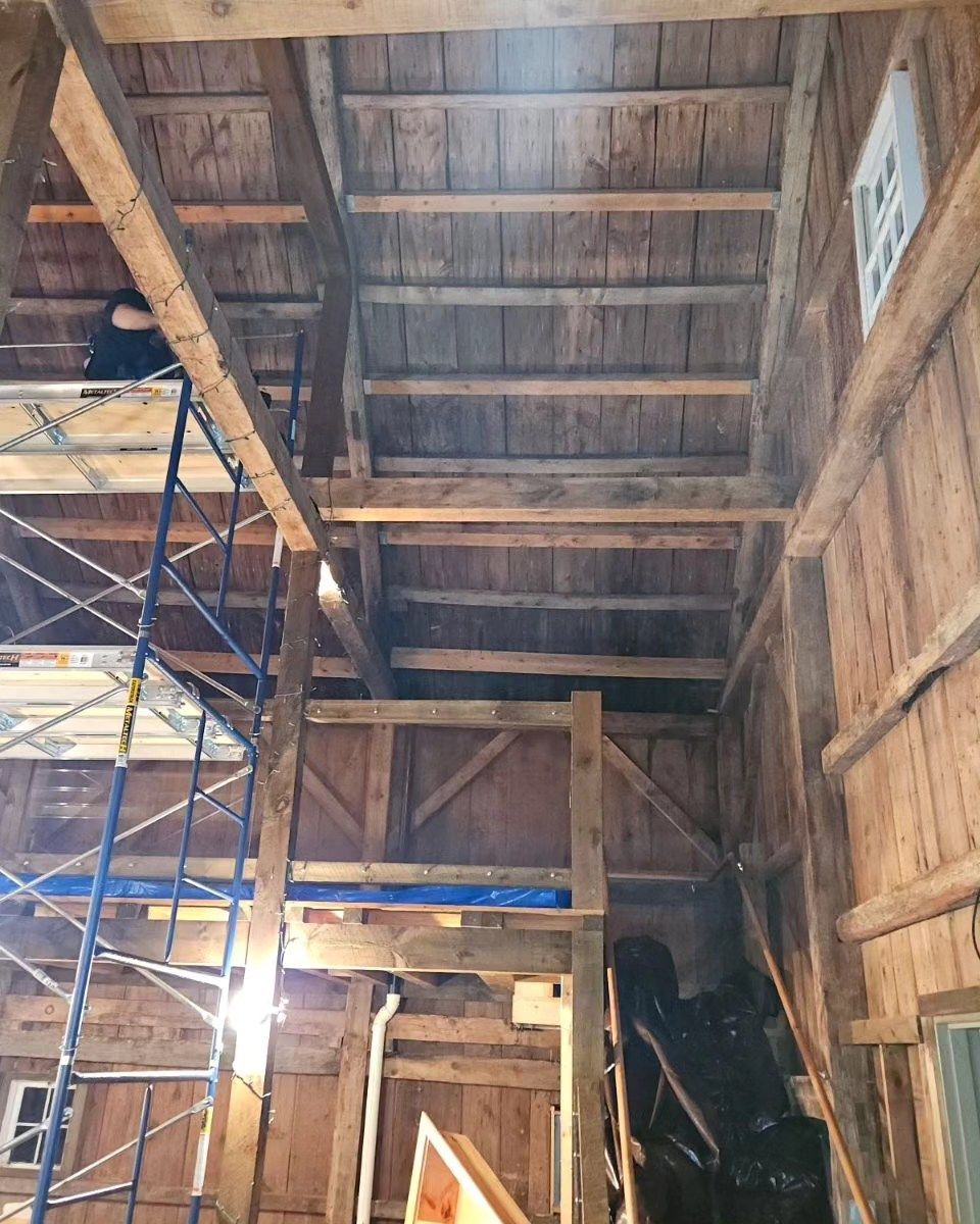 Interior of a rustic barn with wooden beams, scaffolding, and construction materials under a high ceiling.