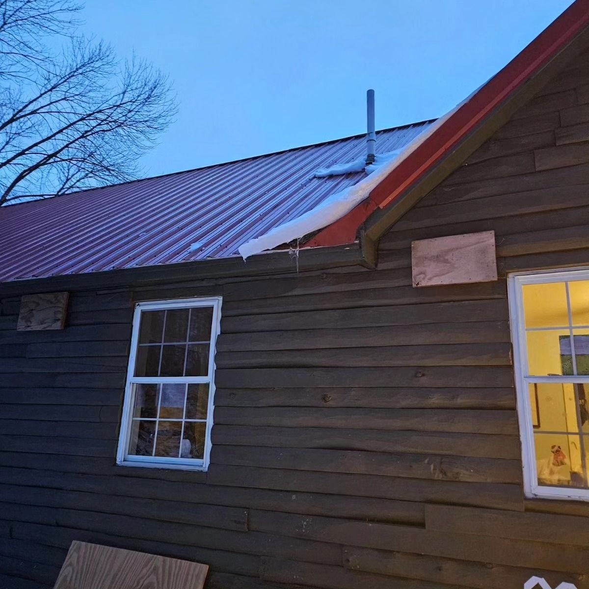 Dark wooden house exterior with red metal roof and lit window at dusk