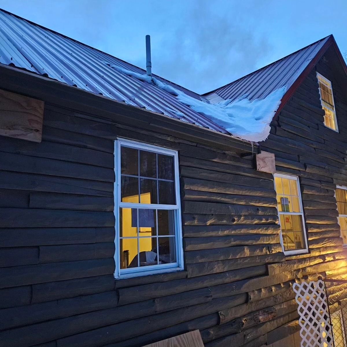 Weathered black wooden house with snow on the roof at dusk, lit windows glowing warm inside.