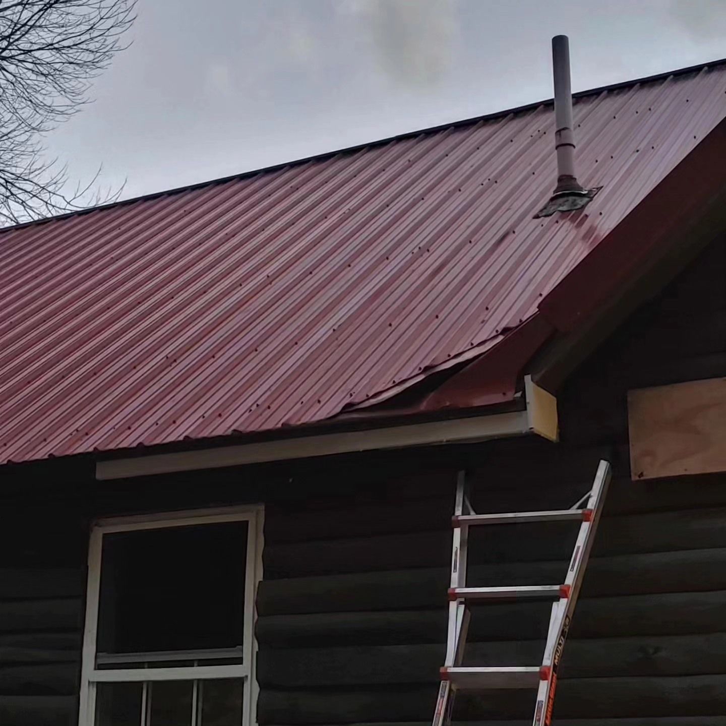 Red metal roof on a dark log cabin with a ladder leaning against the side under a cloudy sky