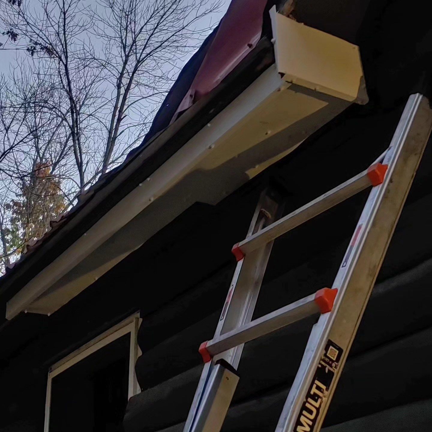 Red-and-white ladder leaning against a dark house roof near a gutter and bare tree branches