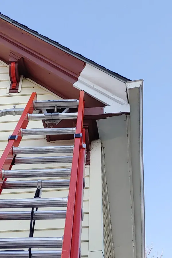 Extension ladder leaning against house roof edge with white gutter and red trim under blue sky