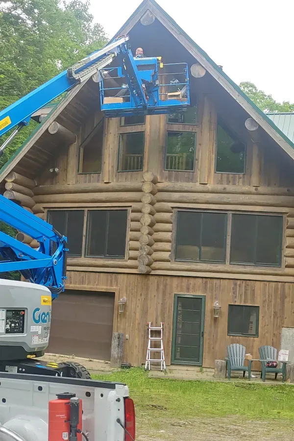 Blue cherry picker lifts workers to the peak of a rustic wooden building with a steep roof.