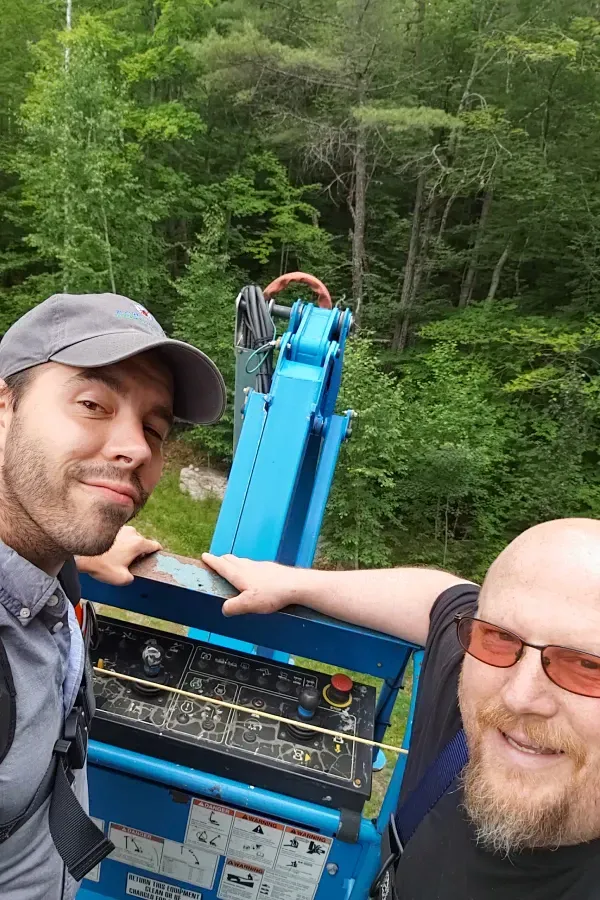 Two men posing beside a blue machine in a wooded area, smiling at the camera.