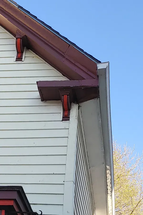 Side of a white house with a brown roof overhang and gray trim against a blue sky