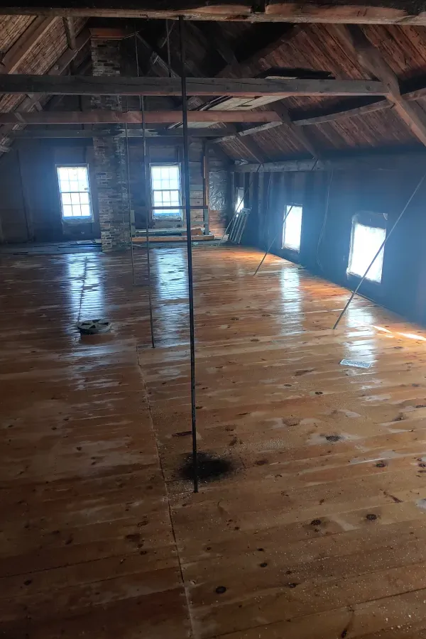 Empty wooden attic with exposed beams, polished floorboards, and sunlight streaming through small windows.