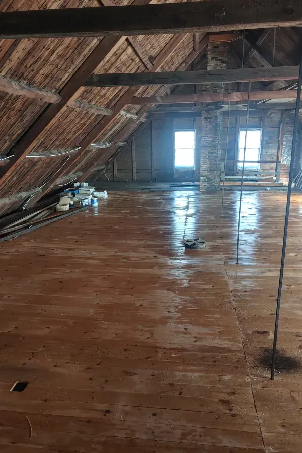 Empty attic with wooden floor, exposed beams, and two bright windows on the far wall.