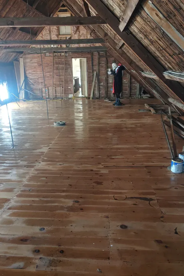Attic interior with wooden beams and a polished plank floor, lit by daylight from a doorway and window openings