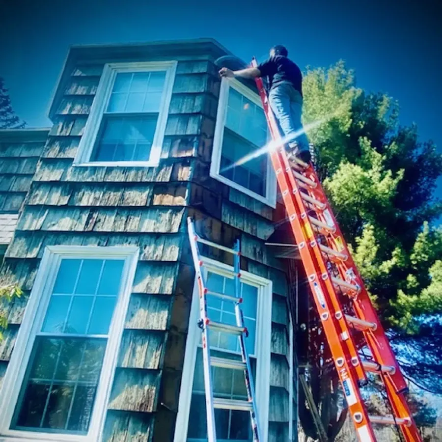 Worker on a ladder repairing a window on a shingled house, with a red extension ladder outside.