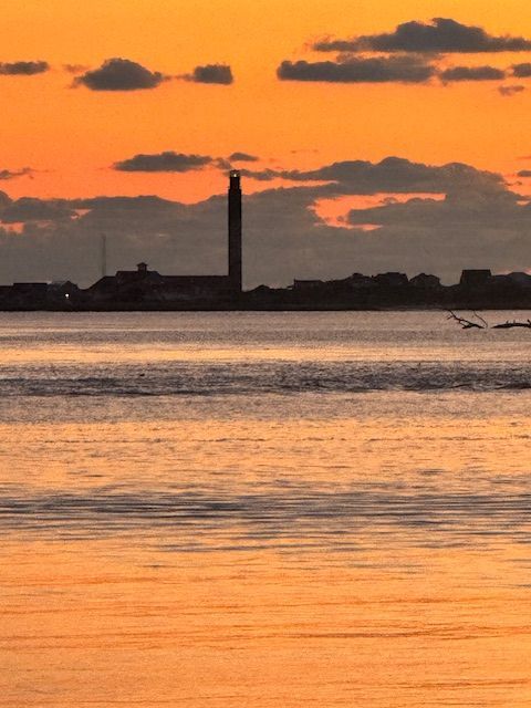 Orange sunset over calm water with dark clouds and a distant shoreline silhouette, including a tall smokestack