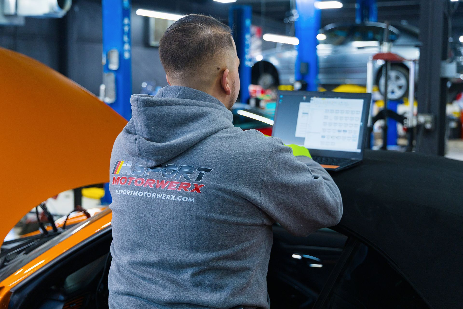 Mechanic at a garage working on a car with a laptop, wearing a grey hoodie. Orange car hood open.