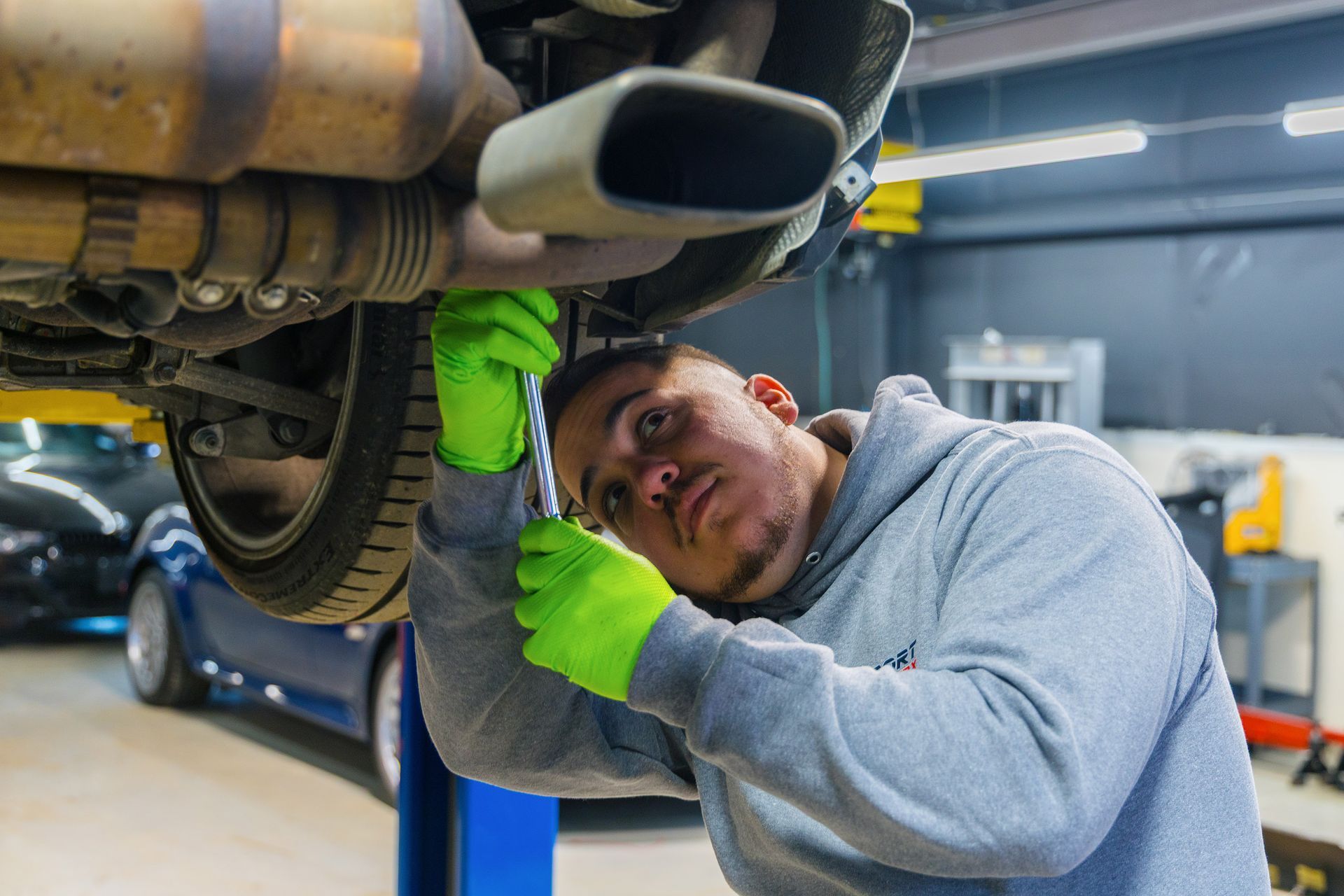 Mechanic in green gloves works under a car in a garage, using a wrench.