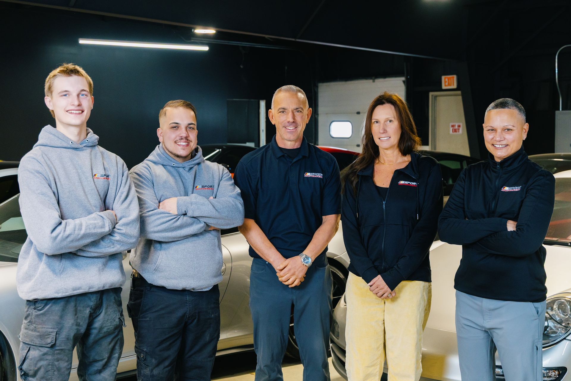 Five people standing in a workshop, arms crossed, in front of a car and equipment area