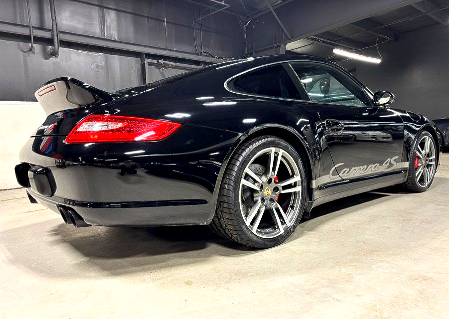 Black Porsche 911 coupe with Carrera side decal in an indoor showroom, rear three-quarter view