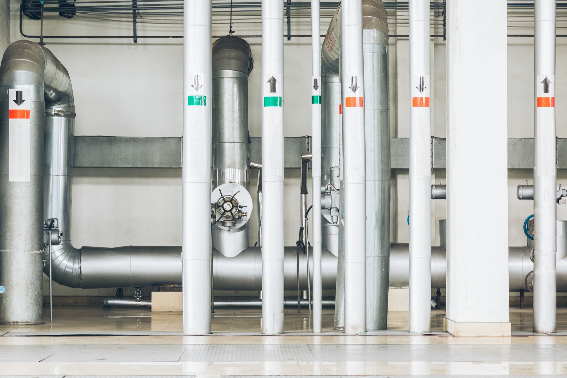 Metallic industrial pipes with color-coded directional flow arrows installed against a white wall.