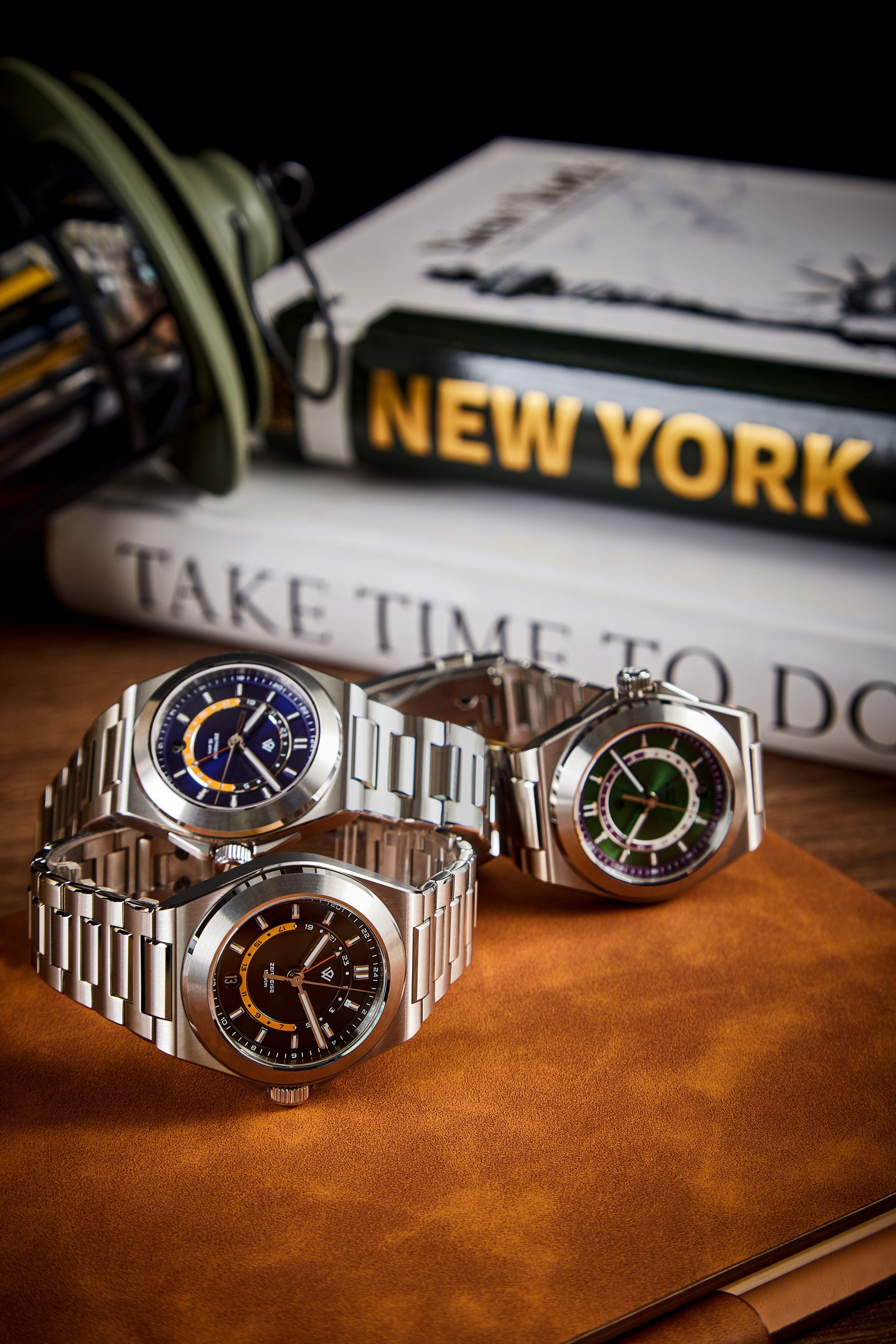Three stainless steel luxury watches with colored dials arranged on a leather surface in front of New York-themed books.