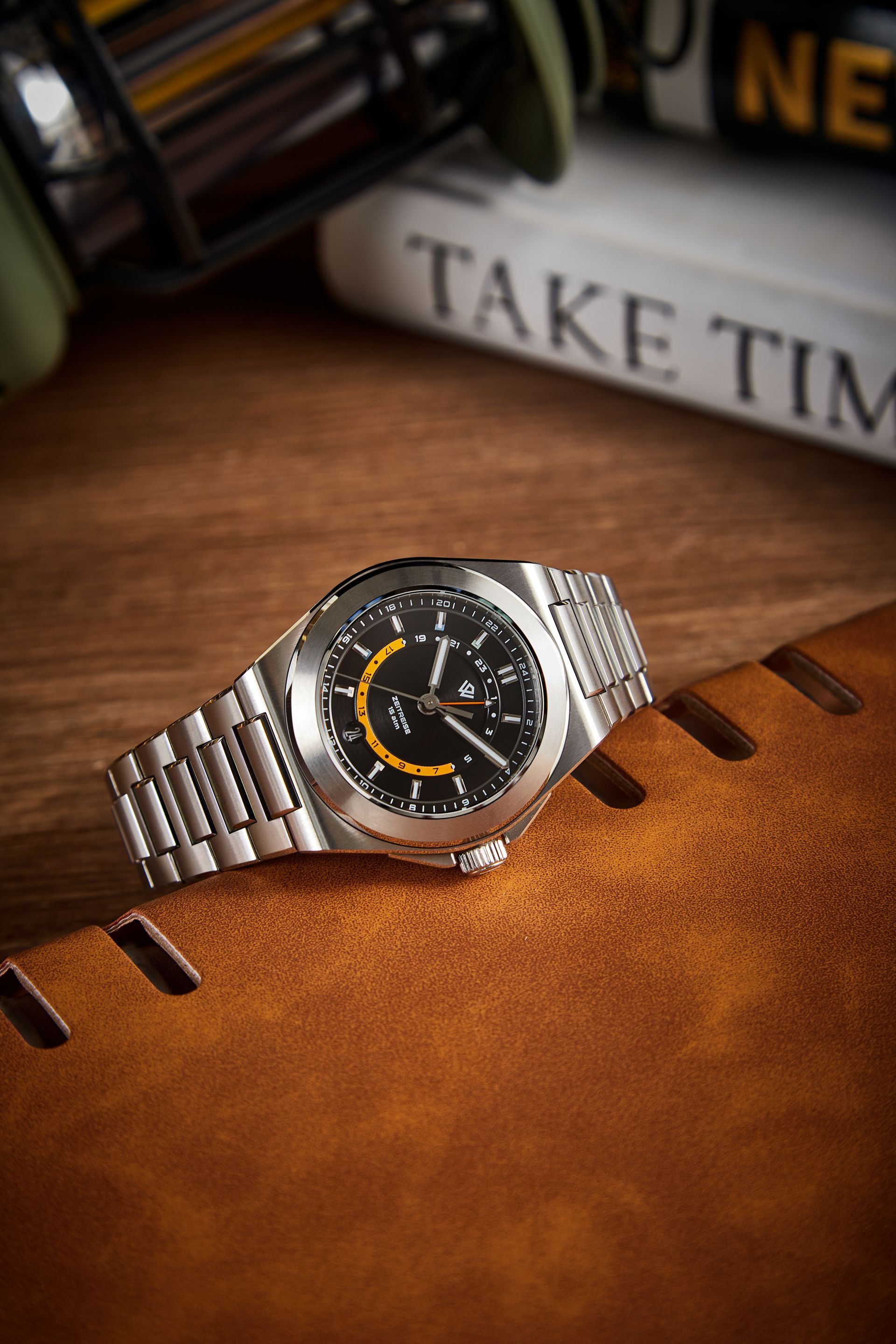A silver metal-link wristwatch with a black and yellow dial sits on a brown leather surface near a book titled 