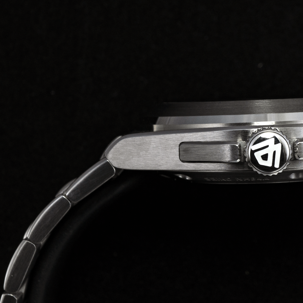 A close-up side view of a silver-toned metal wristwatch with a dark bezel and a logo on the crown against a black background.