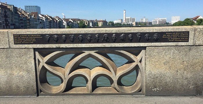 Stone bridge railing with a relief of portrait plaques overlooking a city skyline and river under a blue sky.