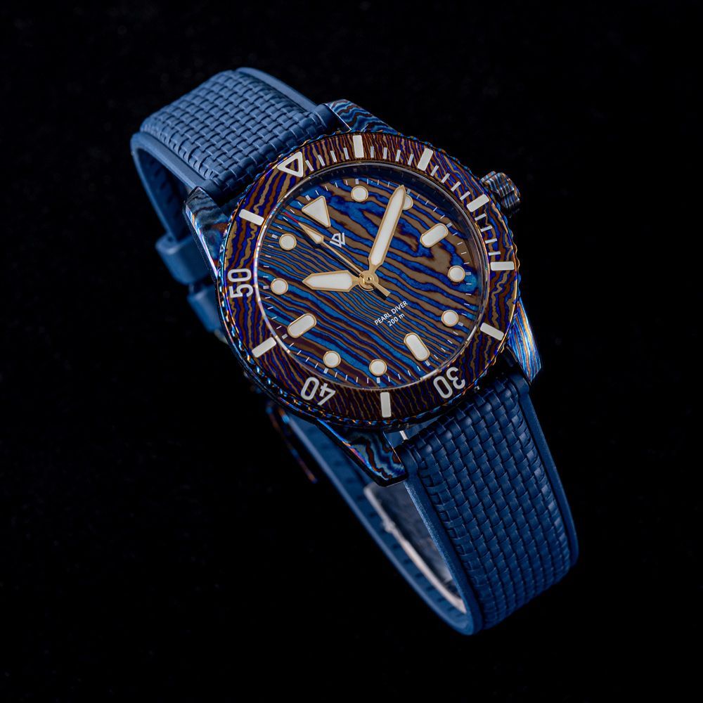 A blue diver watch with a patterned, Damascus-style dial and bezel on a textured rubber strap against a black background.