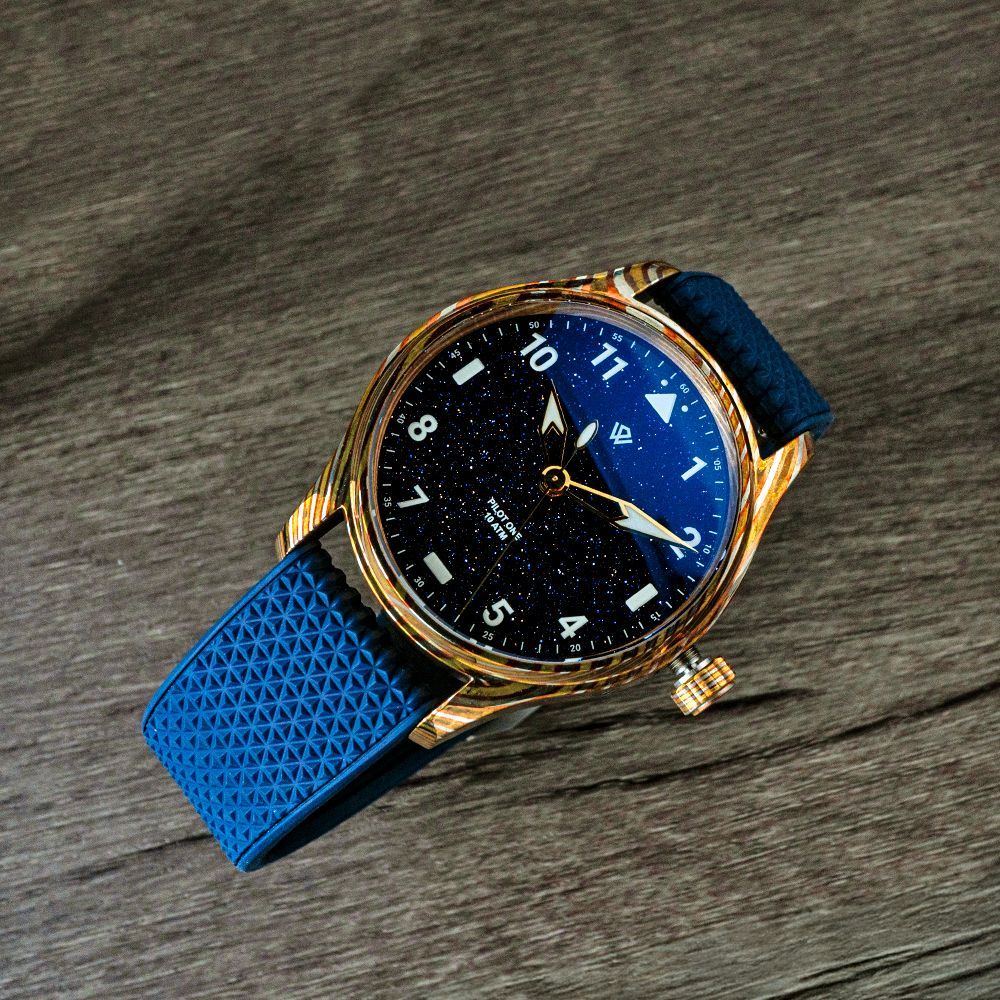 A gold-cased wristwatch with a navy blue starry dial and textured blue rubber strap on a wooden surface.