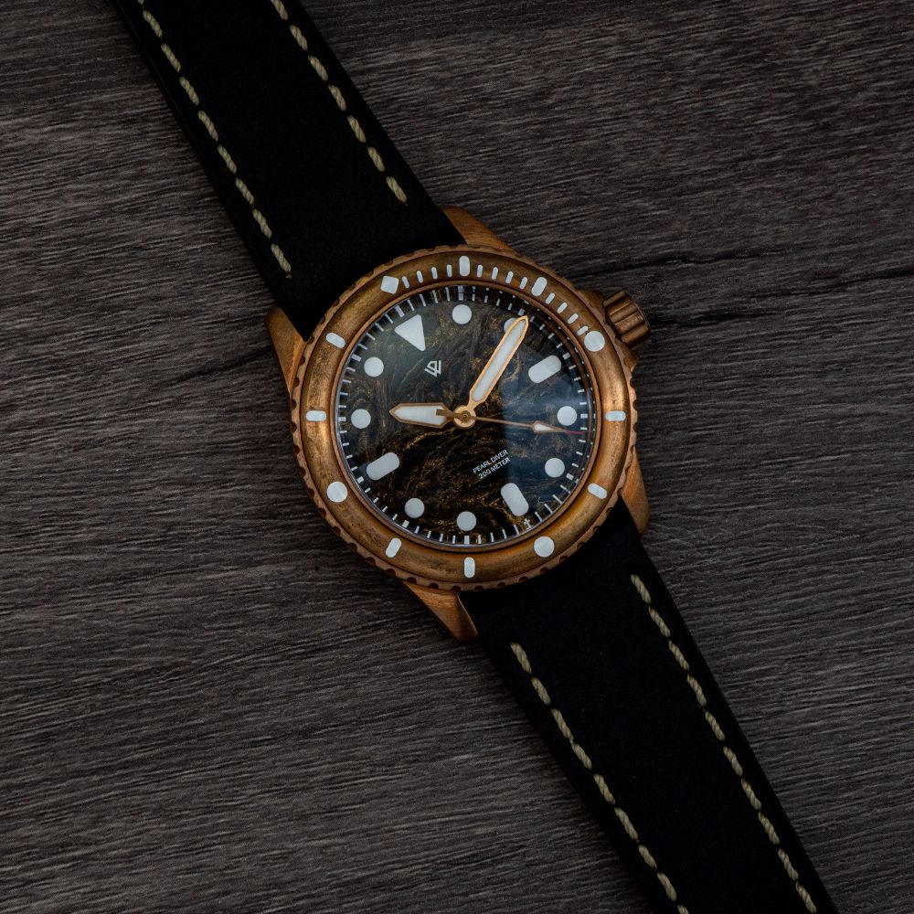 A bronze dive watch with a dark, marbled dial and a black leather strap with white stitching, set on a dark wood surface.