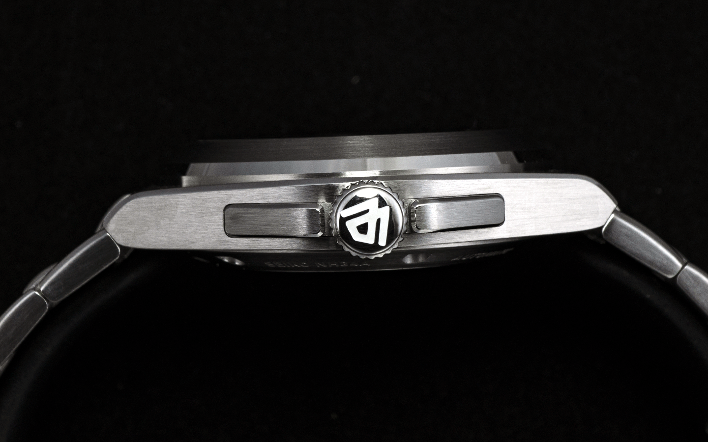 Side profile of a stainless steel wristwatch with a black-and-white crown logo, shown against a solid black background.