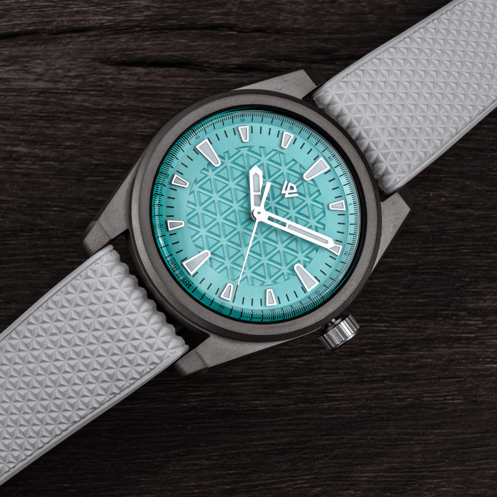 A turquoise watch with a geometric dial pattern and a textured light grey strap, resting on a dark wood surface.