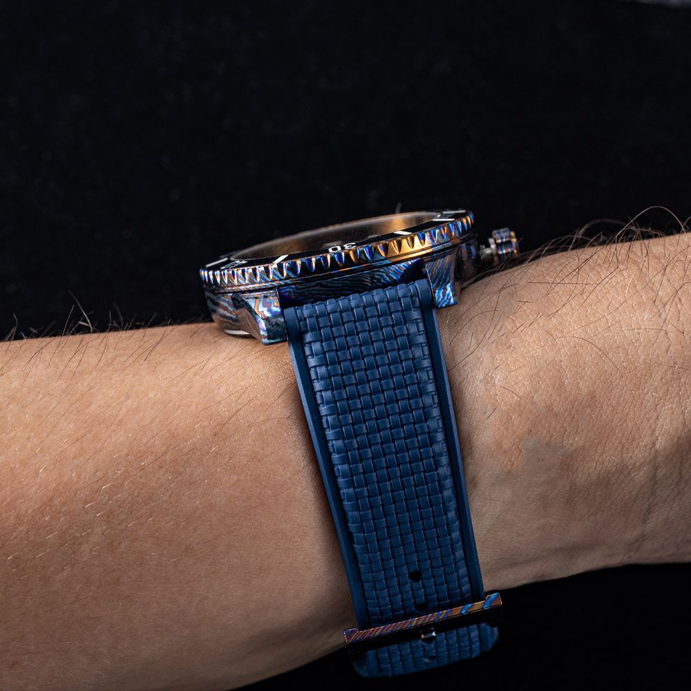 A close-up side view of a wristwatch with a textured dark blue strap worn on a wrist against a black background.