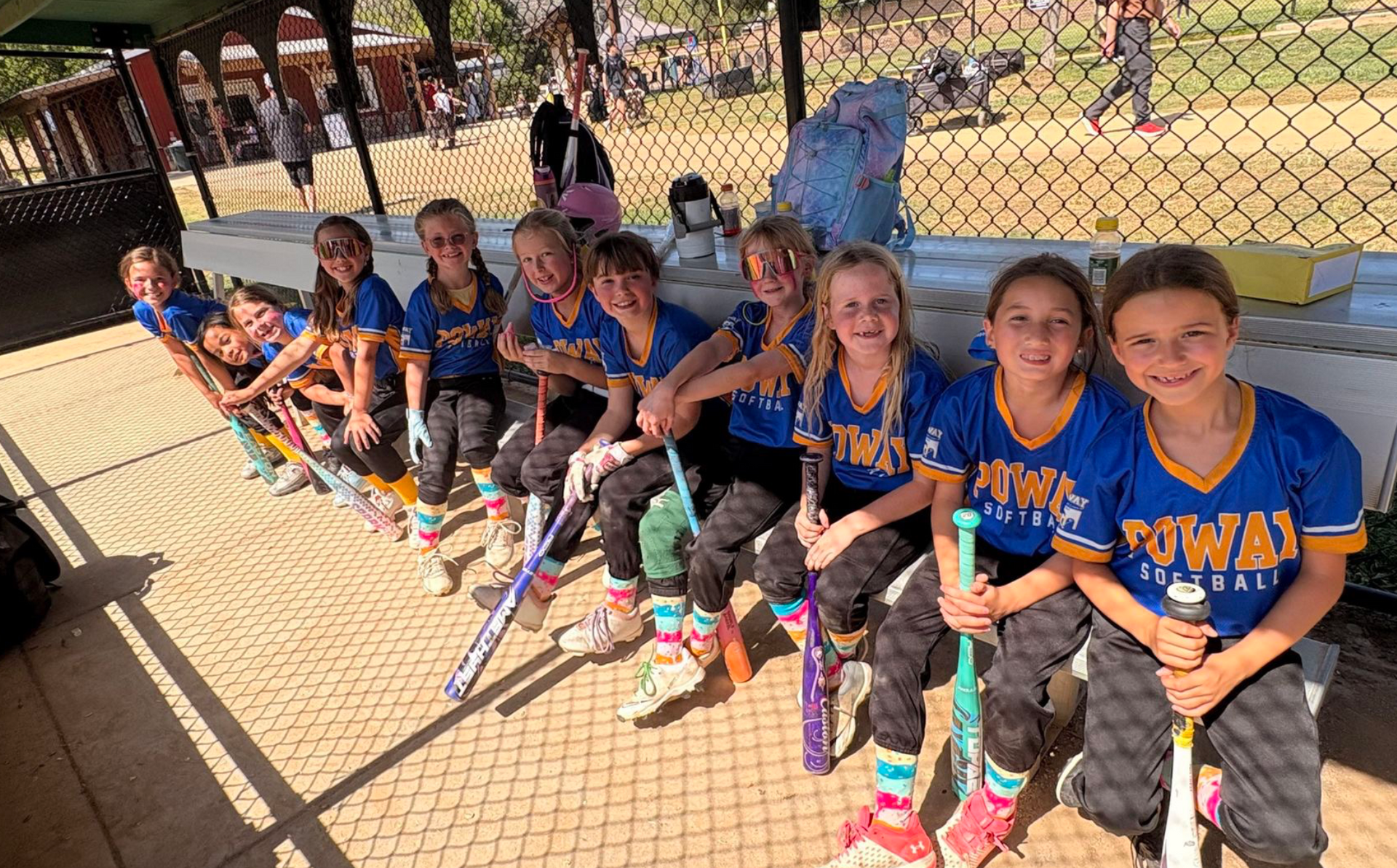 Youth softball team in blue jerseys sitting on a bench behind a chain-link fence at a sunny field.