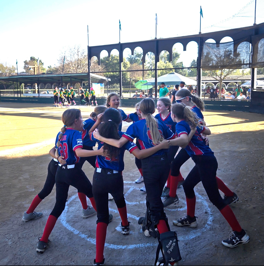 Youth softball team huddling on a field, wearing blue jerseys and red socks, with a dugout in the background.