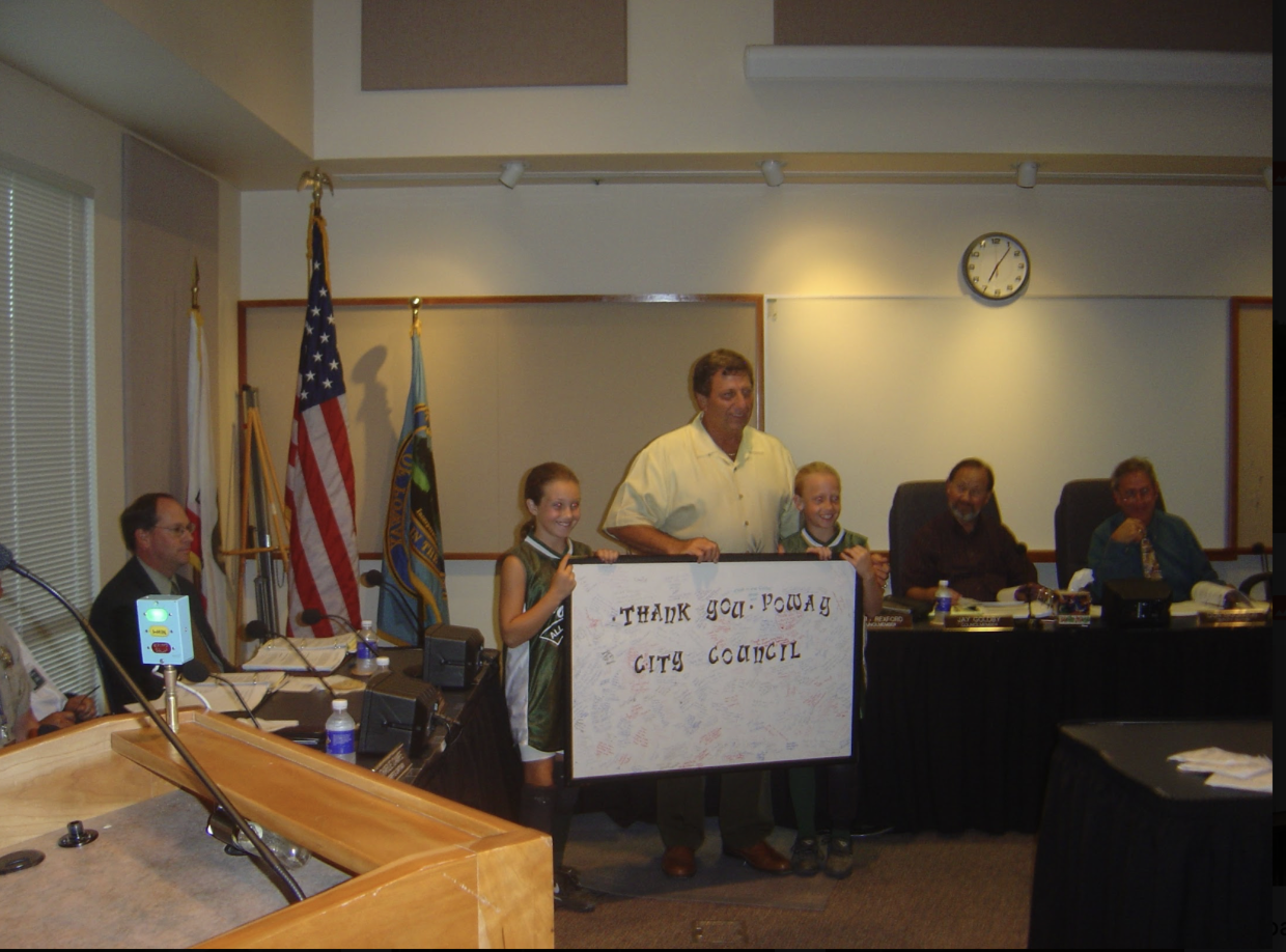 People holding a large sign at a podium in a meeting room with U.S. flags and a wall clock.