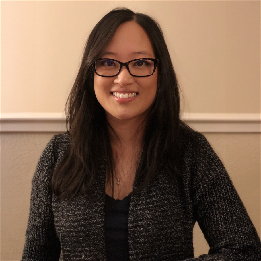 Woman with glasses smiling, wearing a gray sweater, in a room with a light wall.