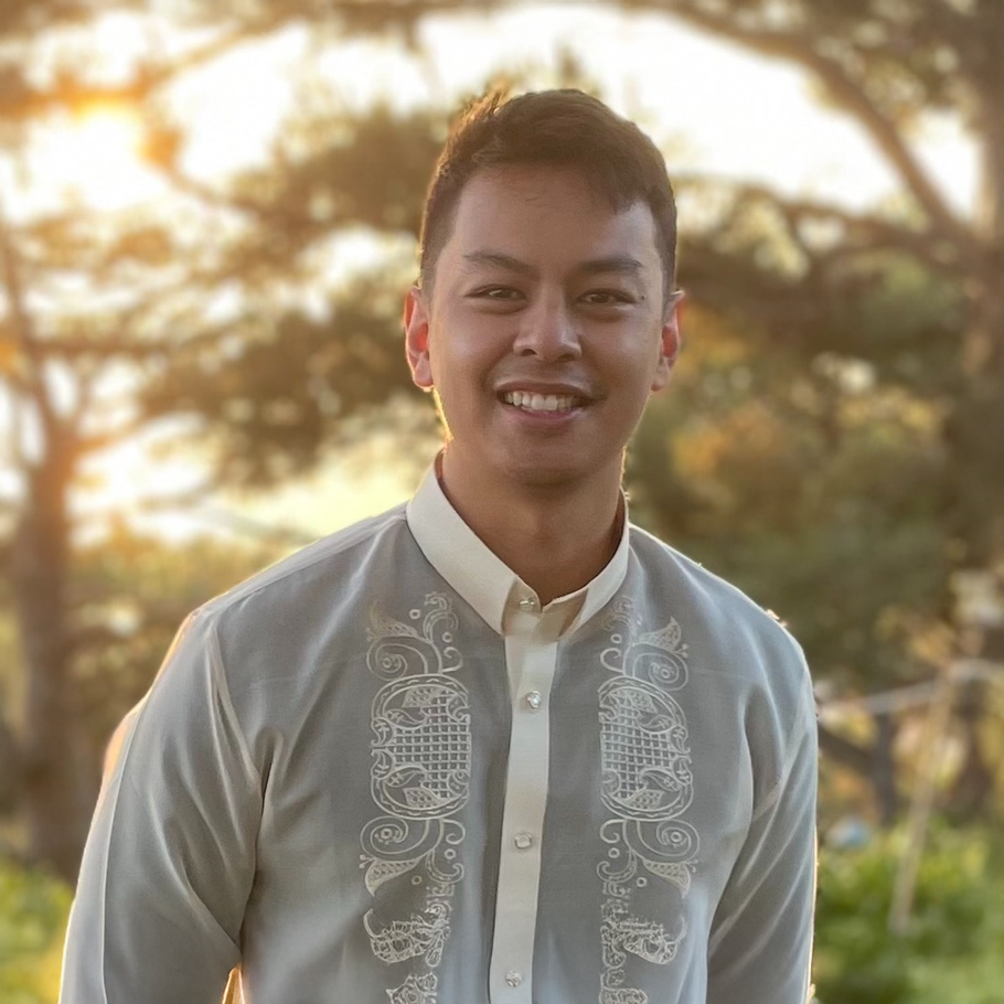 Man in a light-colored embroidered shirt smiles against a blurred outdoor background.