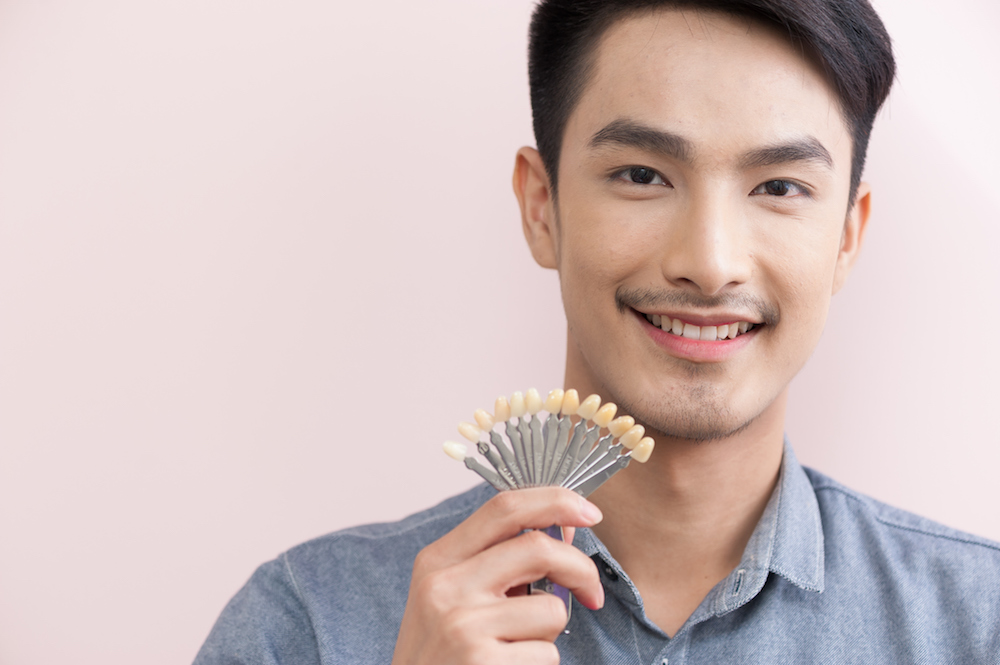 Man holding tooth shade guide, smiling, against a pink background.