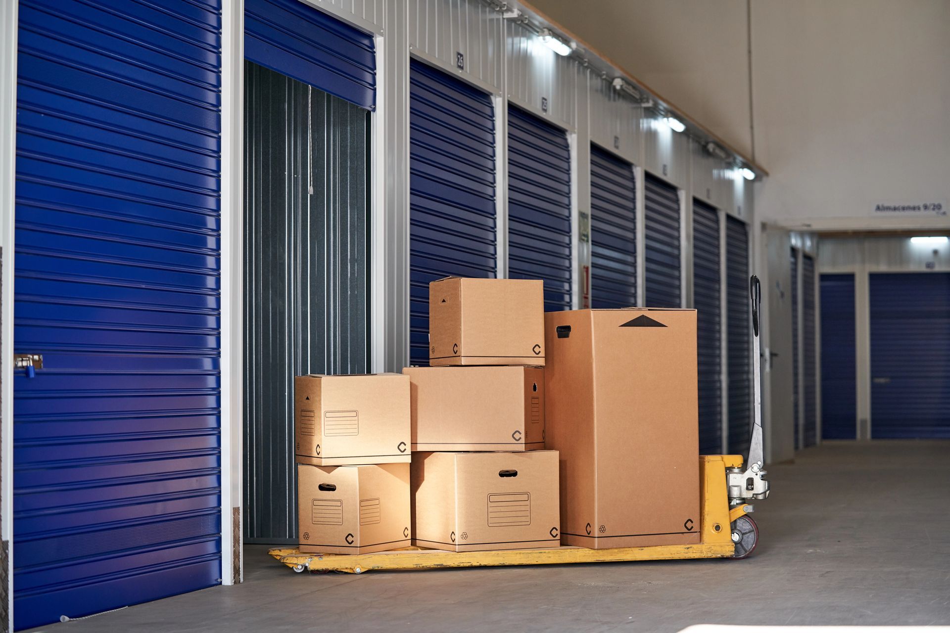 Un transpallet giallo che trasporta scatole di cartone è fermo in un corridoio fiancheggiato da porte blu di depositi self-storage.