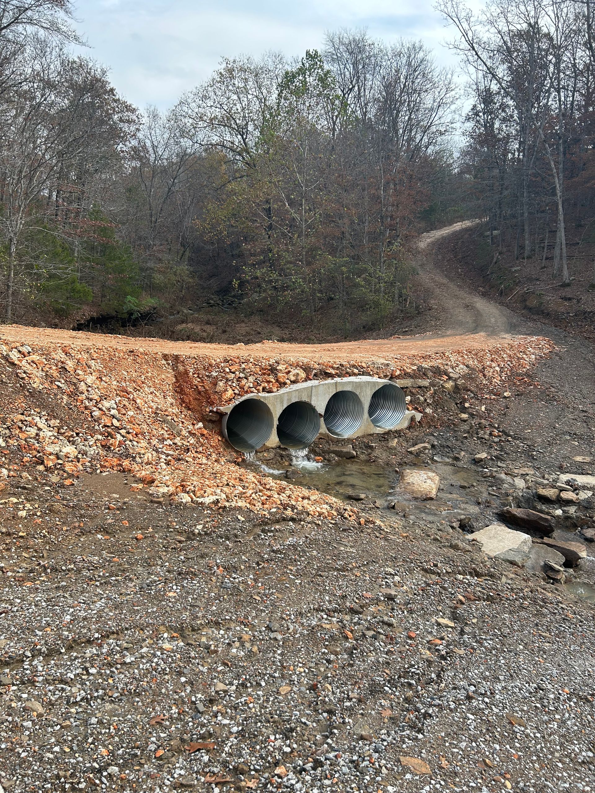A gravel path crossing a creek through concrete culverts, lined with brown stone. Overcast day.